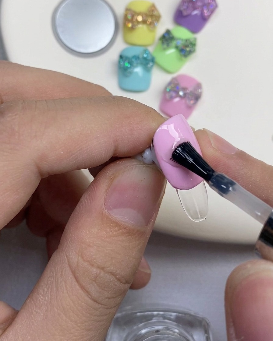A black nail brush applying clear reinforcing gel onto a pink-polished nail.