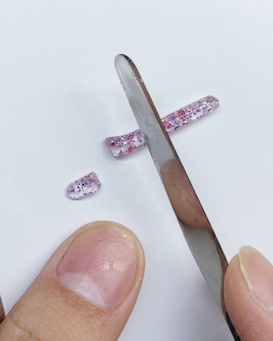 A metal spatula slicing a long strip of glittery pink PVC gel into smaller segments on a white surface.