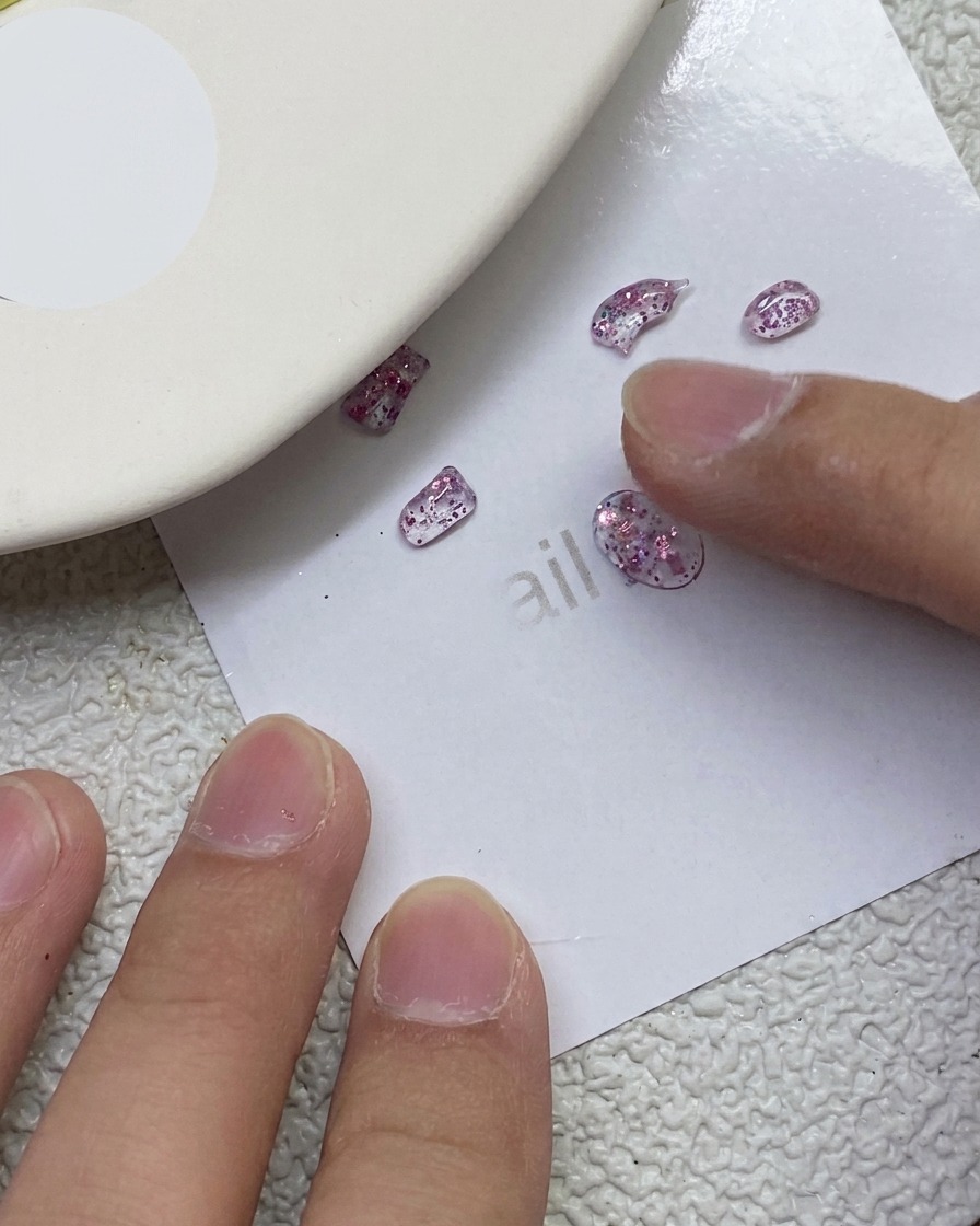 A finger pressing and sculpting small glittery pink PVC gel pieces into bow shapes on a white paper sheet.