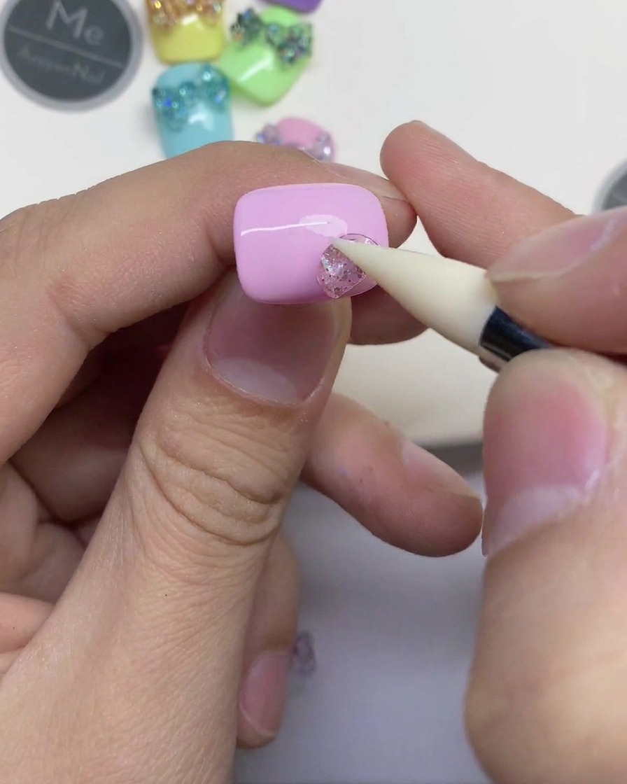 A close-up of a hand using a white silicone tool to press a small piece of clear glittery gel onto a light pink nail.