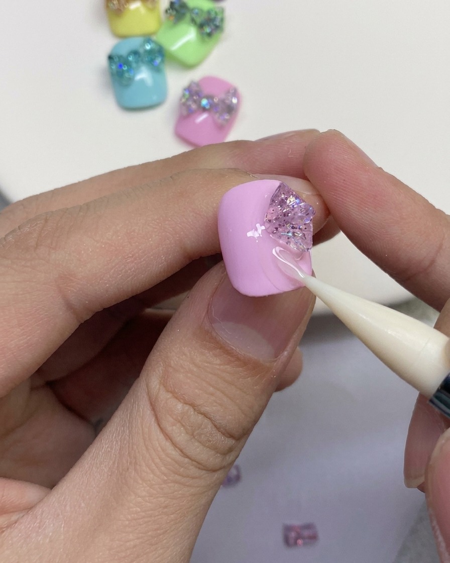 A close-up showing a silicone tool shaping pieces of glitter gel into the outer loops of a butterfly bow on a pink nail.