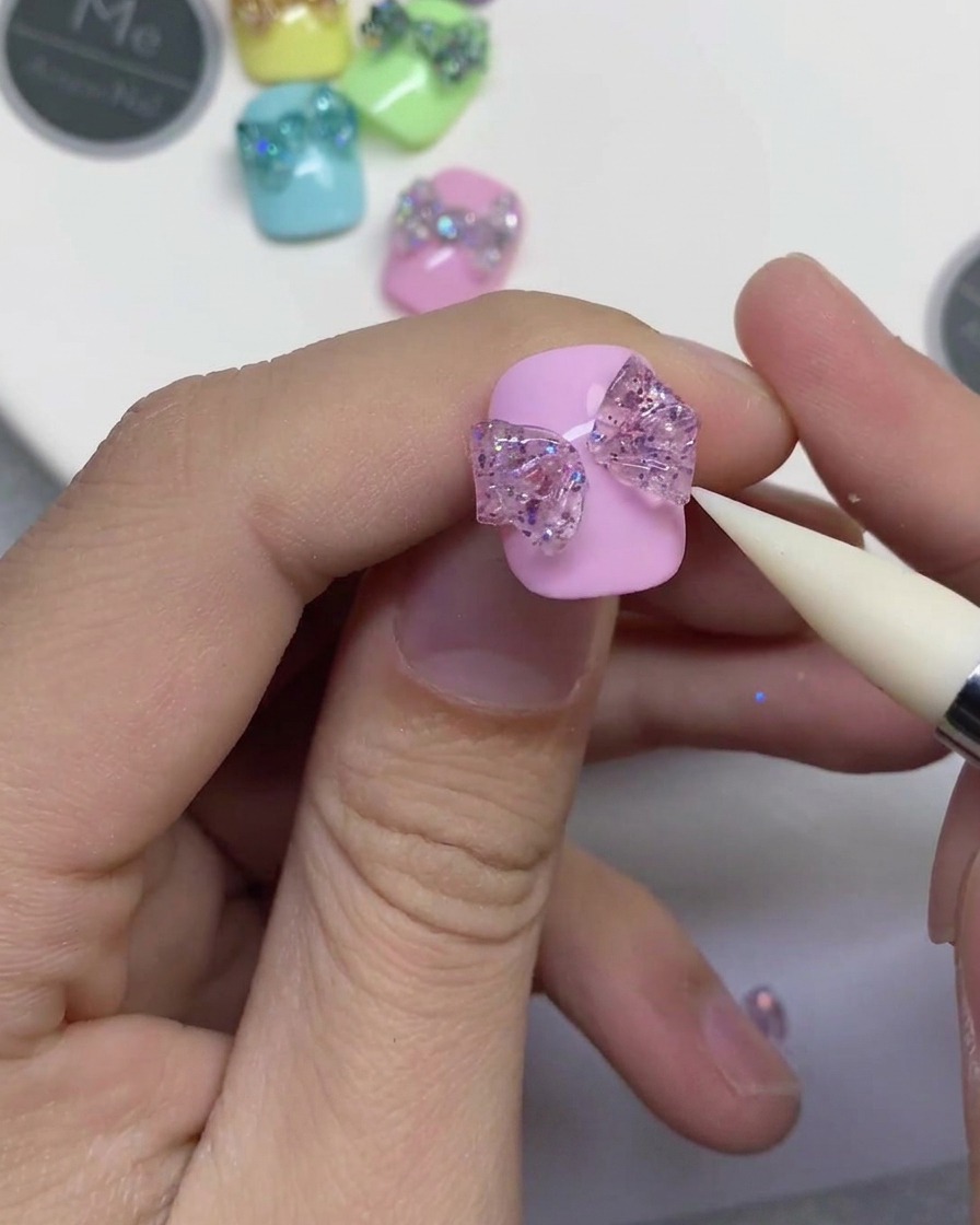A close-up of a white silicone tool pressing into a center strip of glitter gel to connect two bow loops on a pink nail.