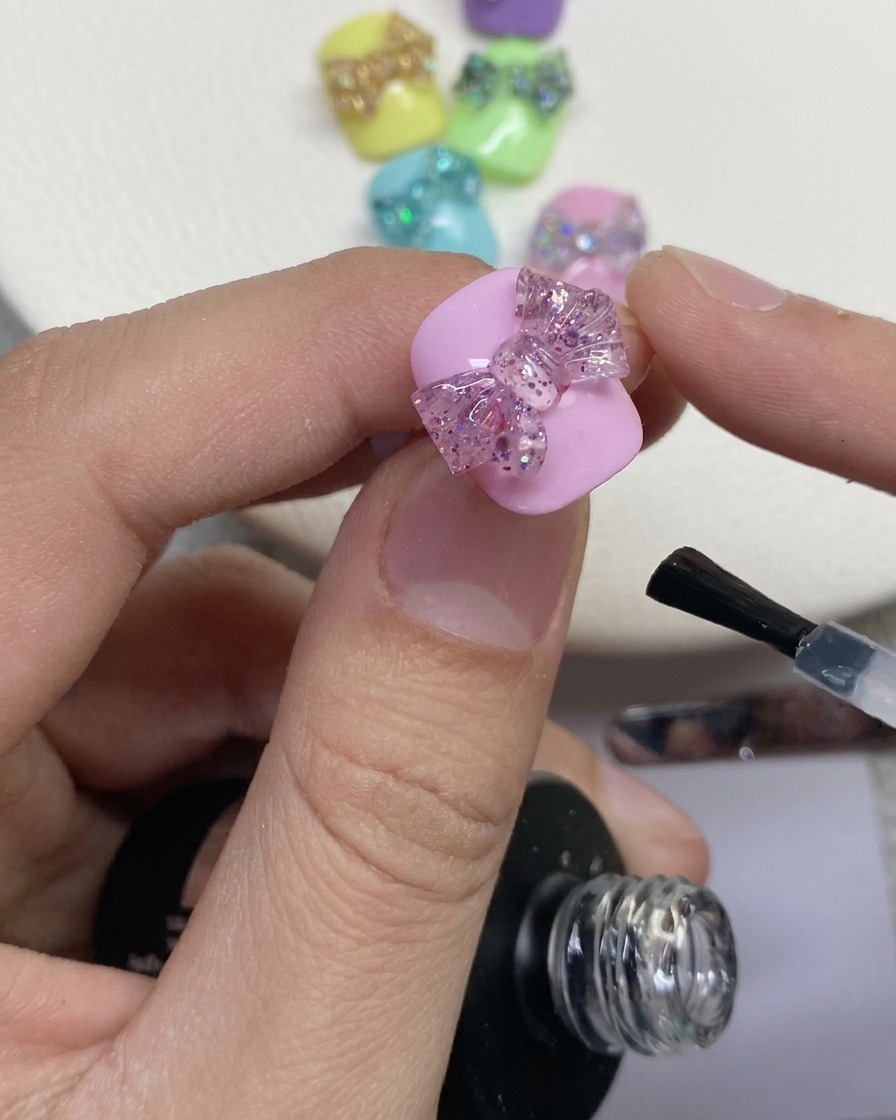 A close-up of a nail brush applying clear top coat around a 3D glitter bow on a painted pink nail.