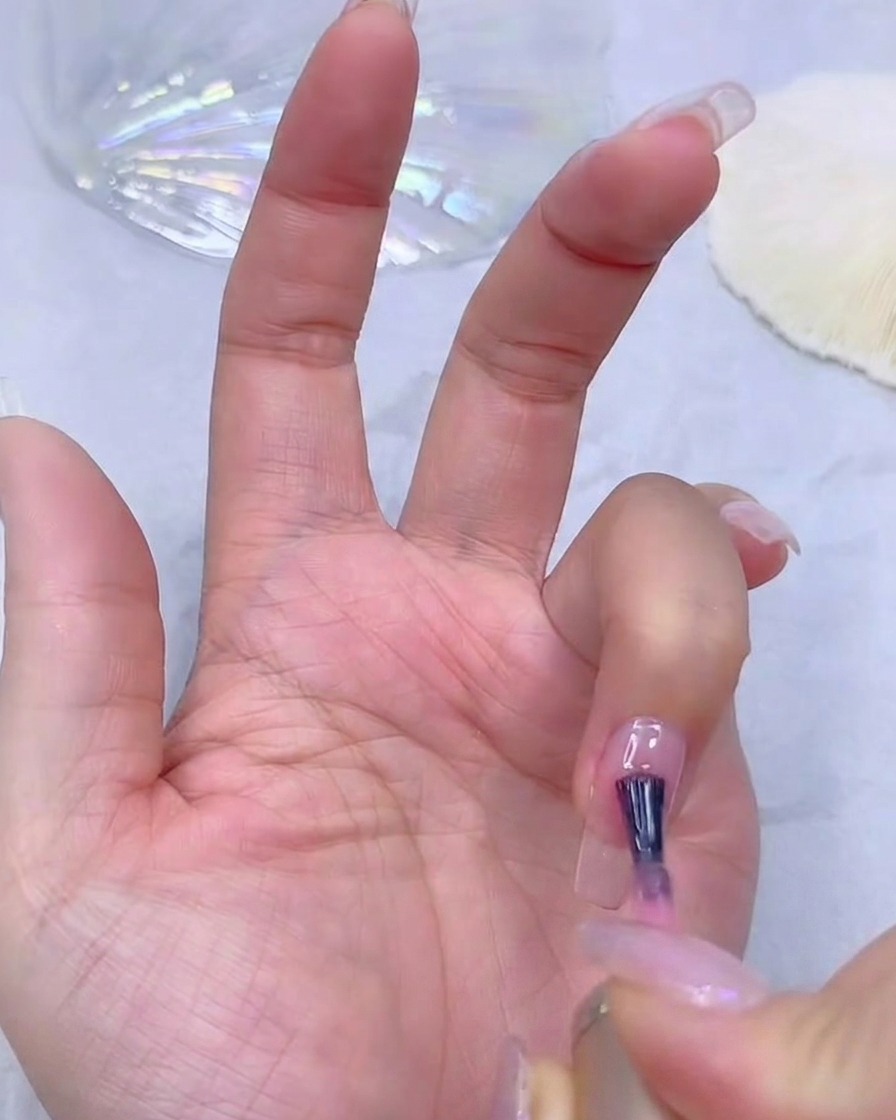 A nail technicians hand applying a sheer pink gel base coat to a clear nail extension.