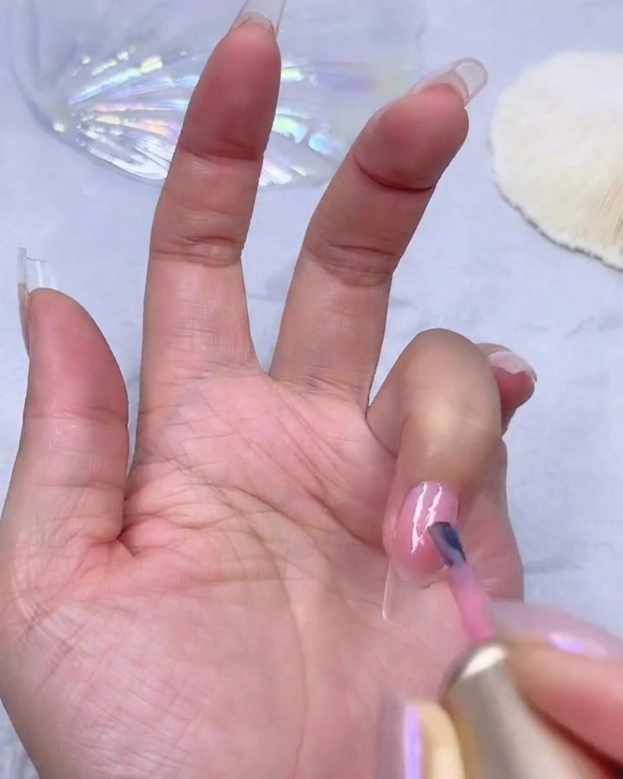 Applying a second coat of sheer pink gel polish to build color opacity on a nail.