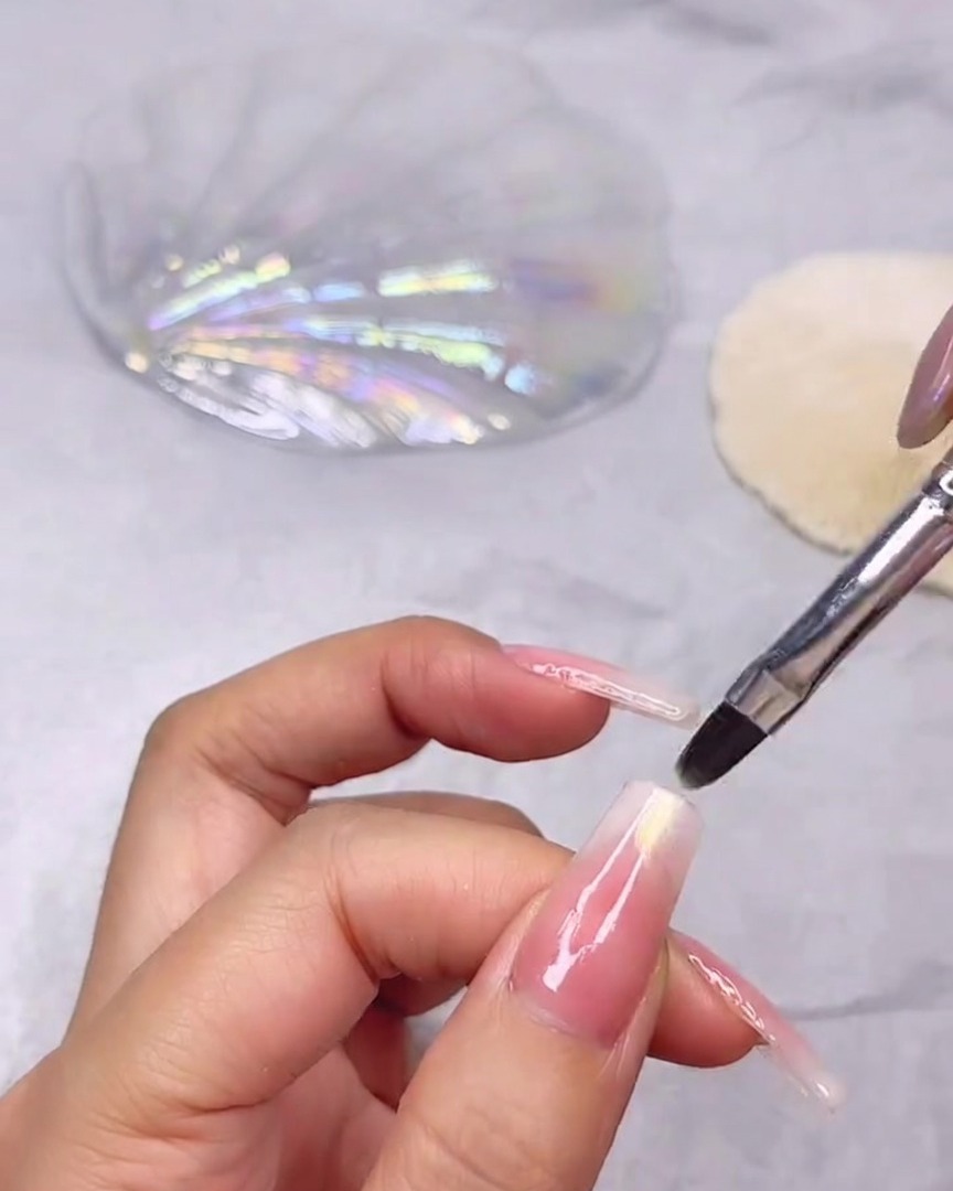 A fine nail brush applying shimmering clear shell gel to the tip of a long nail with a pink and white gradient.