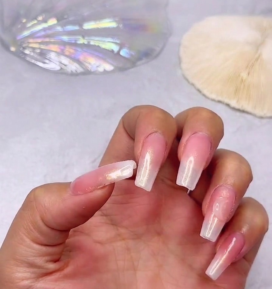 A close up of a hand displaying long glossy nails with a pink and milky white gradient featuring a large clear heart rhinestone placed near the thumb cuticlecite: 1.