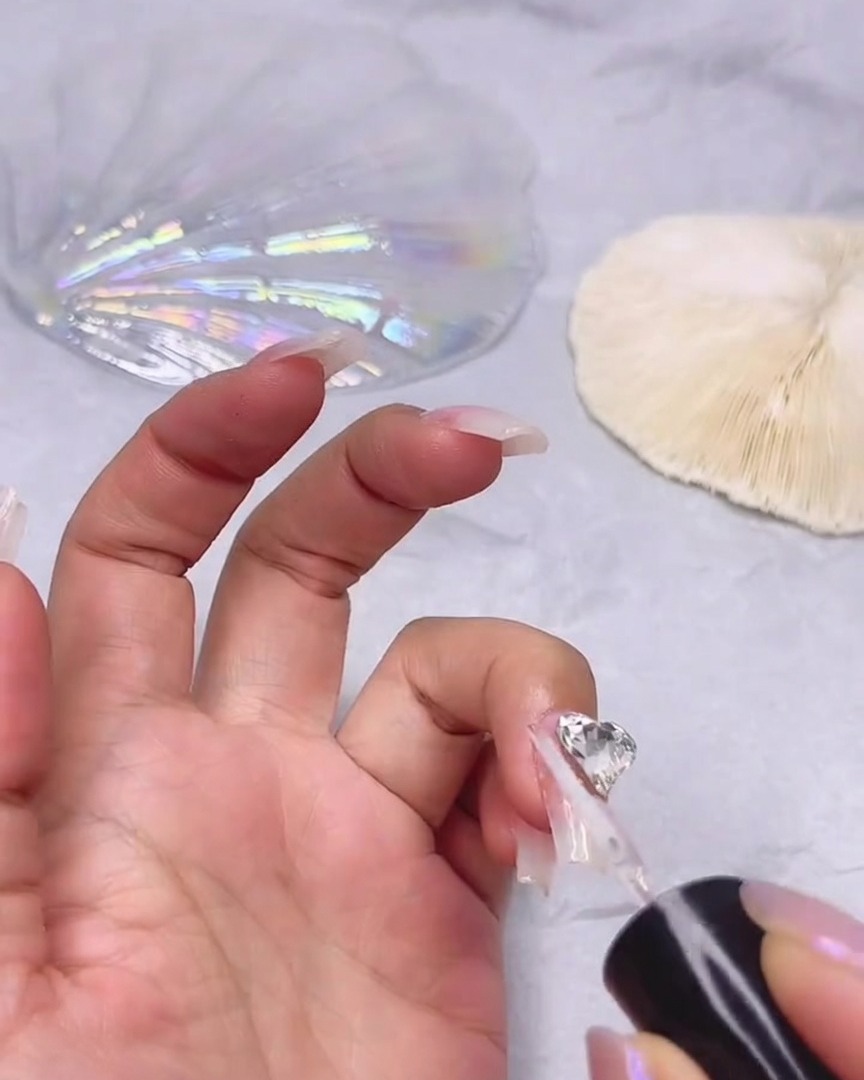 A fine brush applying clear sealing gel around the edges of a large heart-shaped clear rhinestone attached to a fingernail.