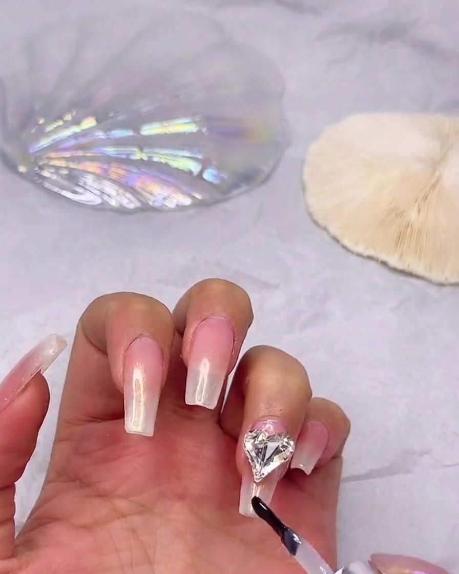 A gel polish brush applying shiny top coat over a gradient nail, carefully working around a secured heart rhinestone.