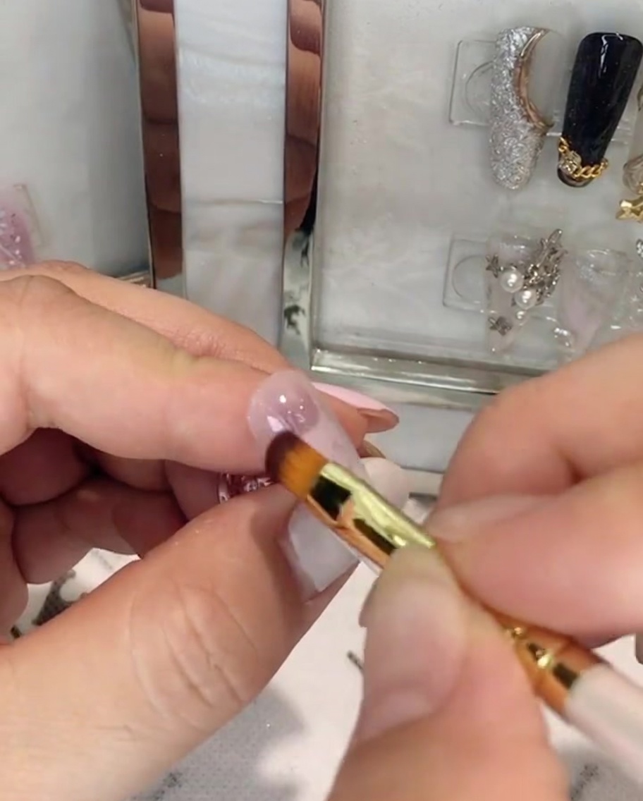 A hand holding a nail brush applying pink gel to the upper half of an almond-shaped nail.