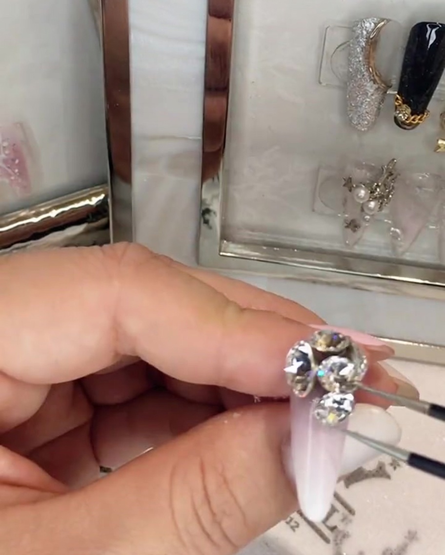 Pointed tweezers arranging small rhinestones into a large cluster on the fingernail.