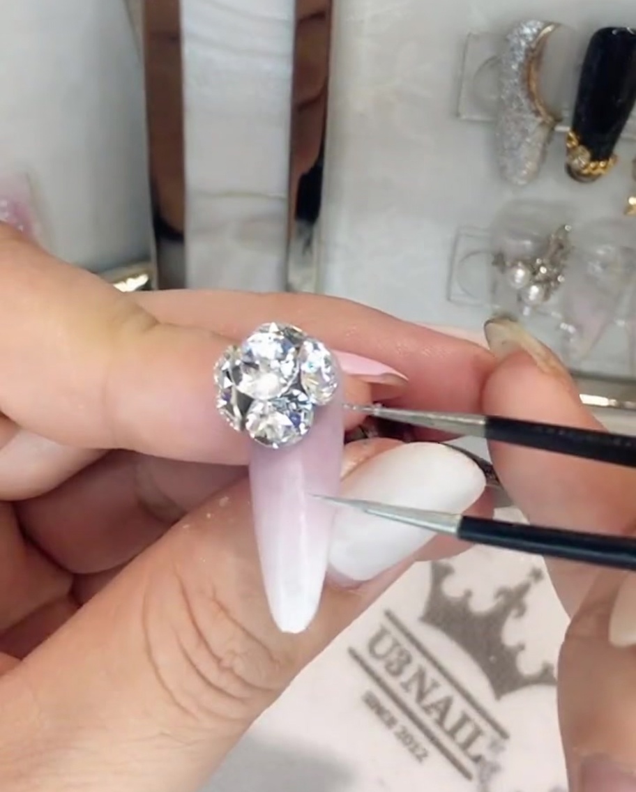 A pair of precision tweezers adjusting individual crystals in a large rhinestone cluster on a nail.