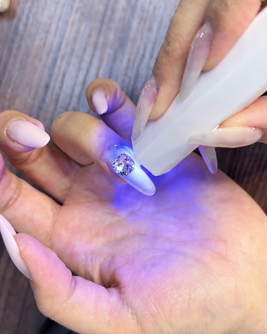 A close-up of a hand picking up small crystal rhinestones to arrange them around a larger center stone on a manicured nail.