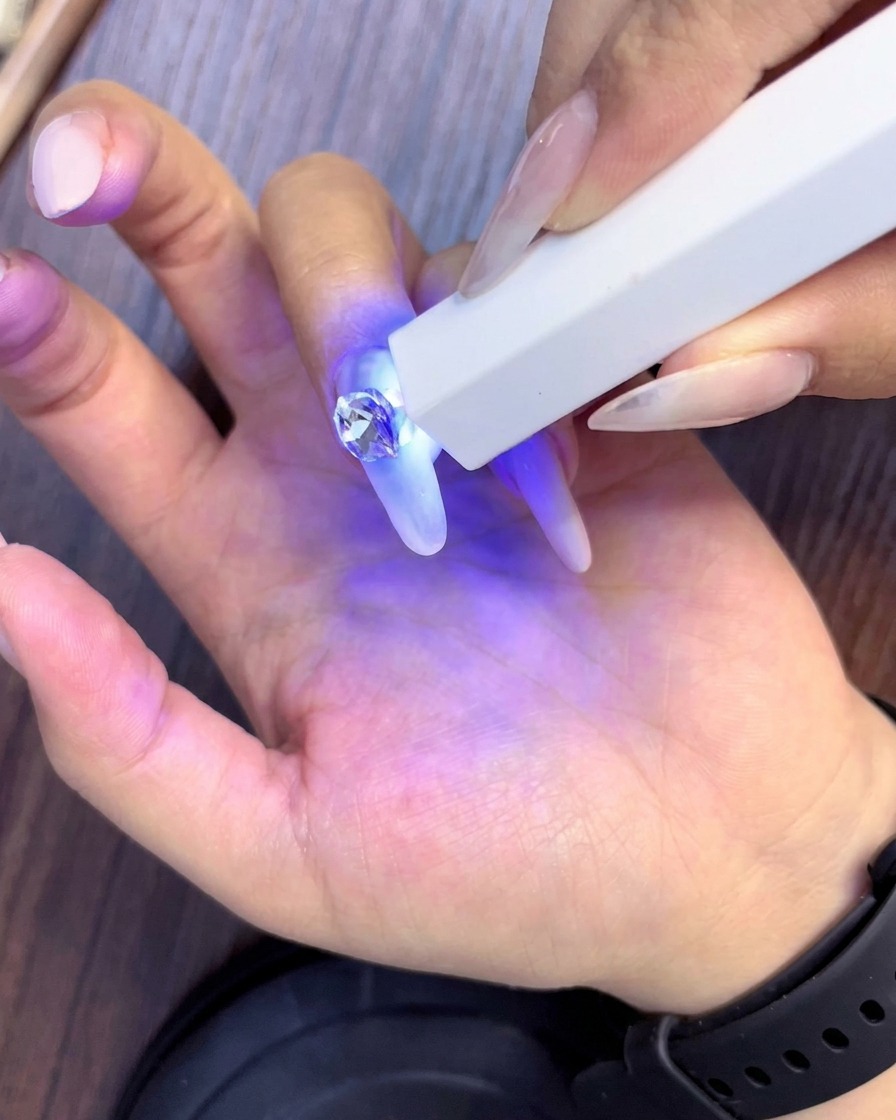 A handheld UV lamp curing newly placed rhinestones surrounding the main center crystal on an almond-shaped nail.
