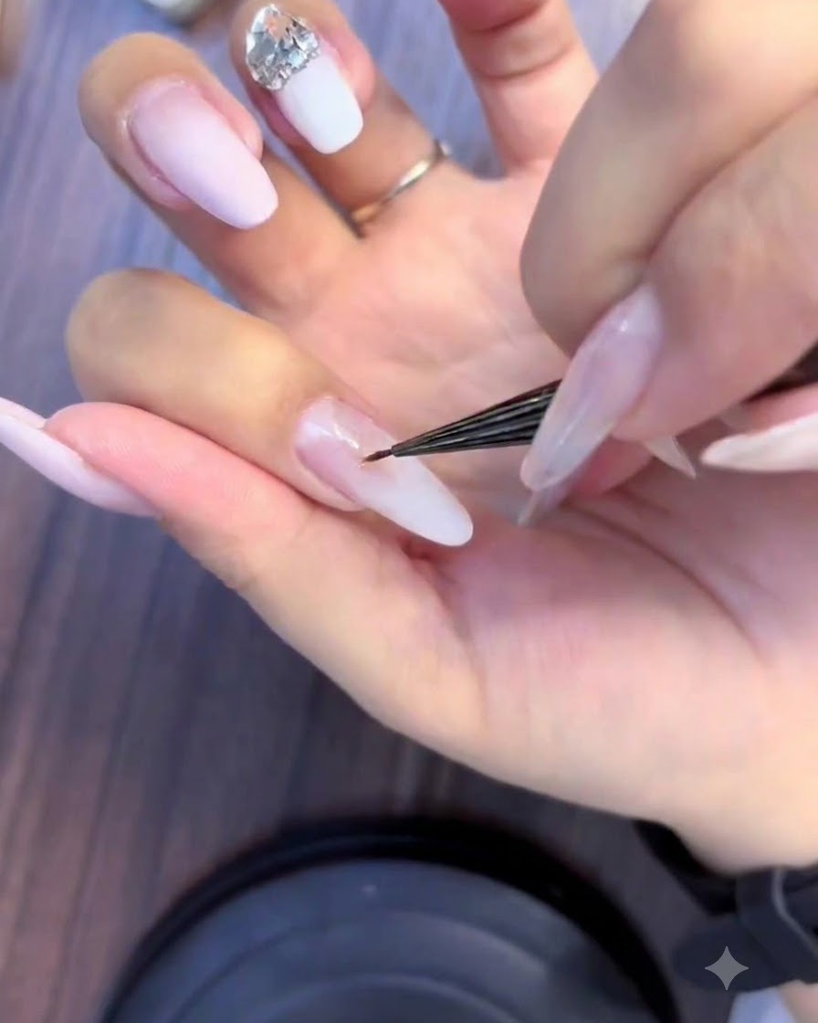 A close-up of a nail technician using a fine brush to apply small rhinestones into a crystal cluster on a pink and white ombre nail.