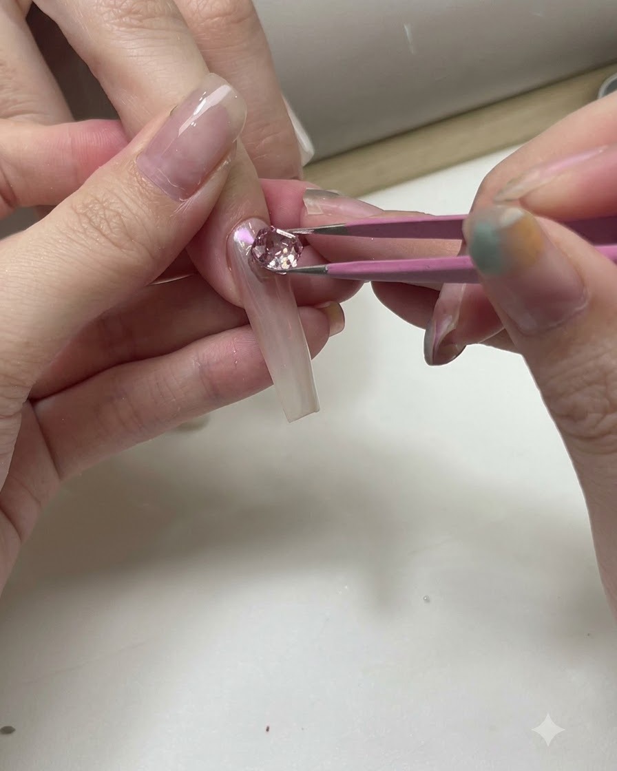 A pair of tweezers positioning sparkling pink and clear crystals onto a transparent practice nail tip.