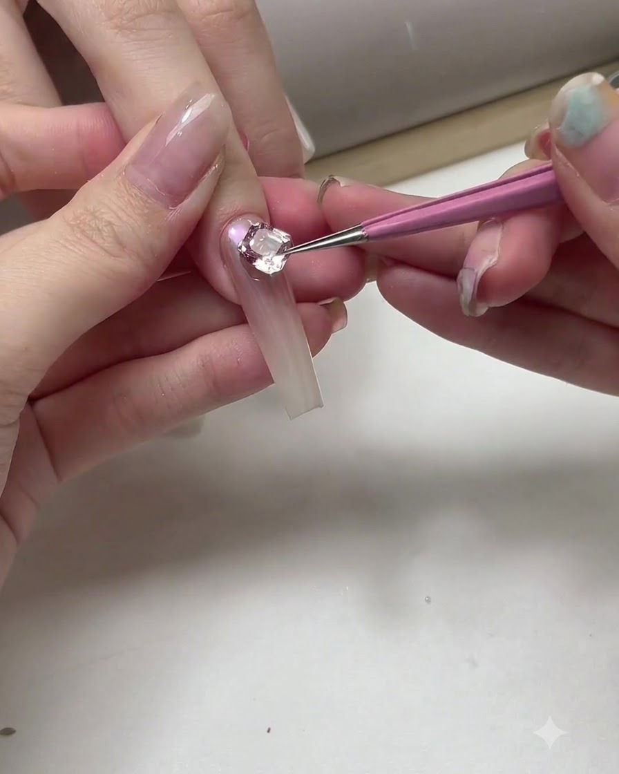A close-up of a manicurist using a thin brush and silver gel to add delicate curved outlines near a large crystal on a nail.