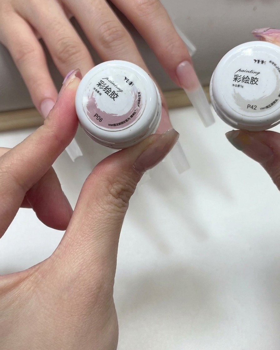 Hands holding two small pots of pink and white painting gel with extended nails visible in the background.