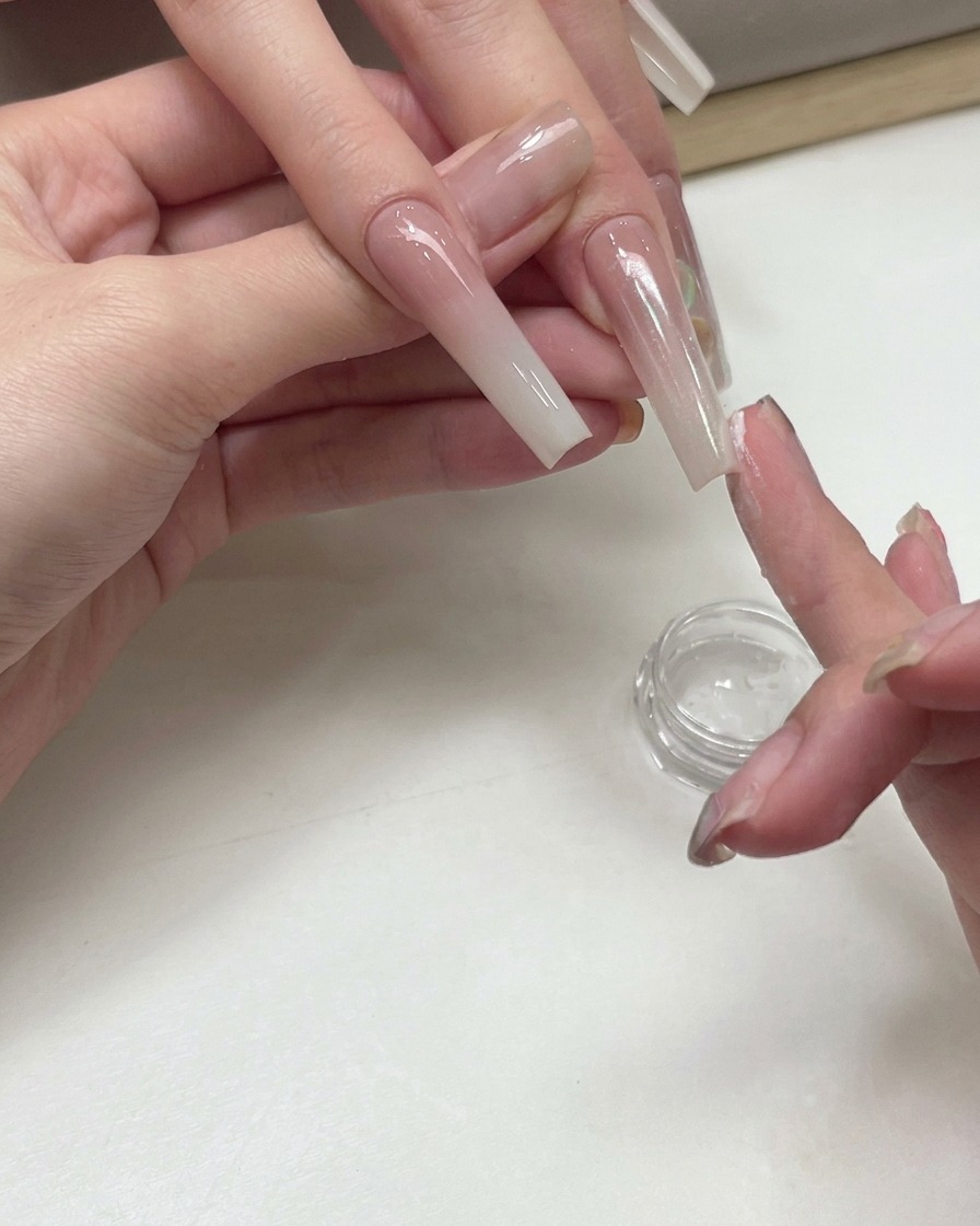 A manicurist using a fingertip to pat shimmering white peach pigment powder onto the surface of an extended gradient nail.