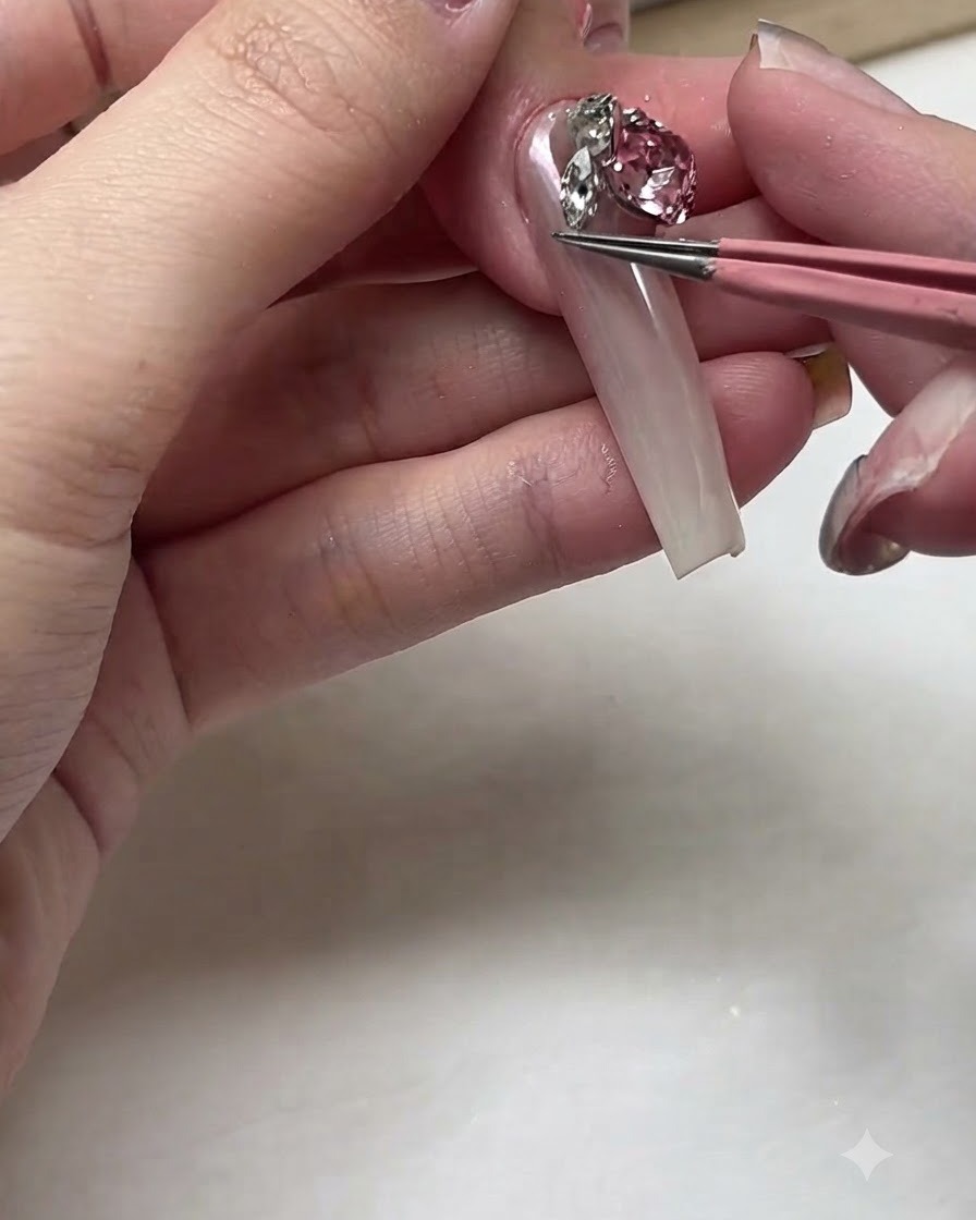 A pair of pink tweezers placing a large sparkling pink crystal rhinestone onto a long nail bed.