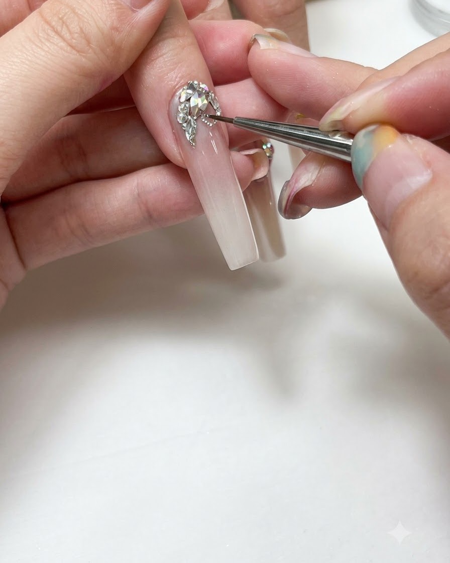 A close-up of a manicurist using a fine detailing brush to paint silver gel lines around clear rhinestones on a long, ombre extended nail.