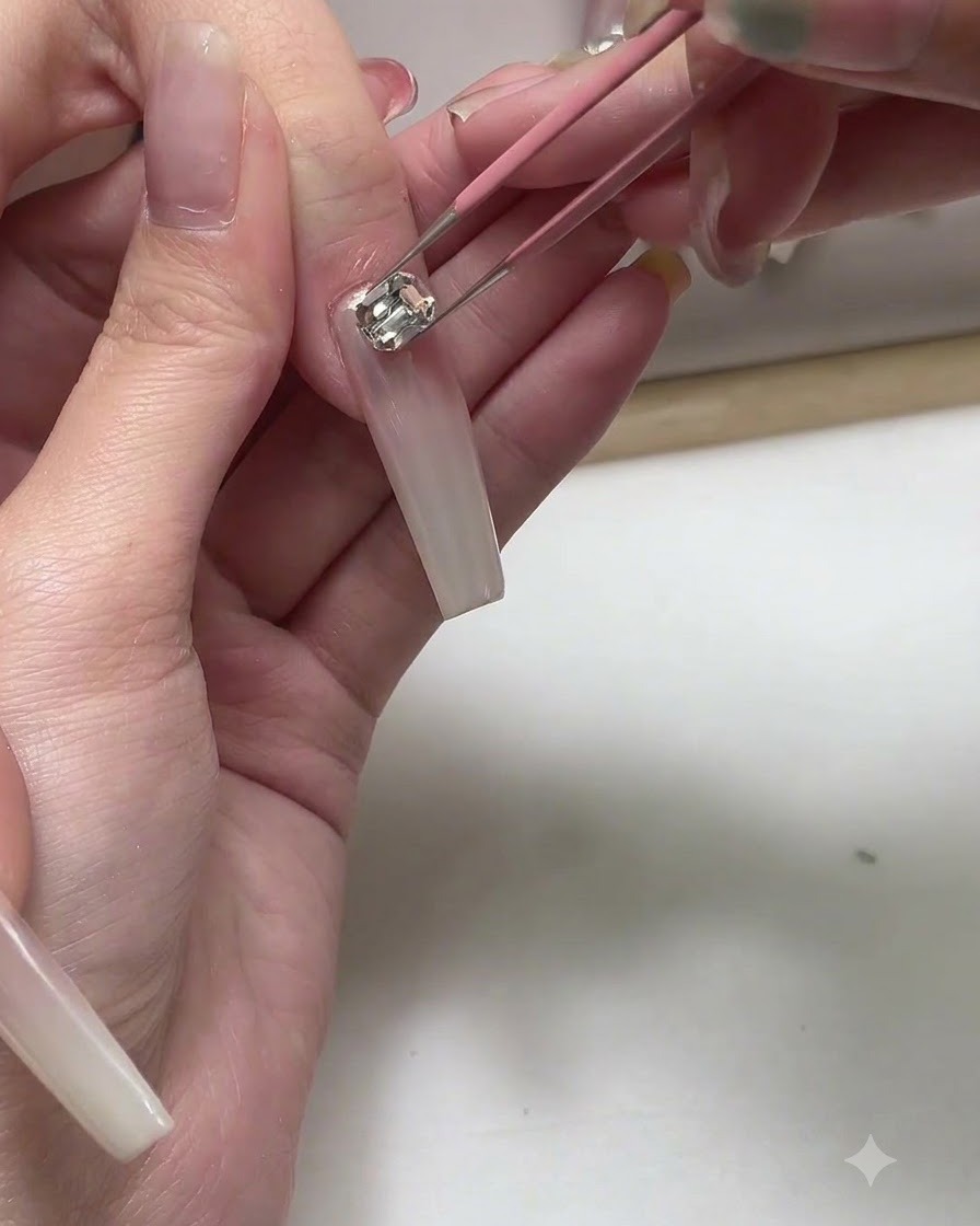 A pair of tweezers placing a small metallic charm onto a long nude-pink gel nail.