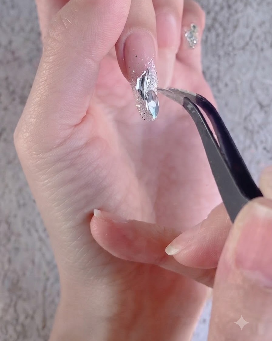 A hand using tweezers to arrange white irregular rhinestones on a decorated nail.