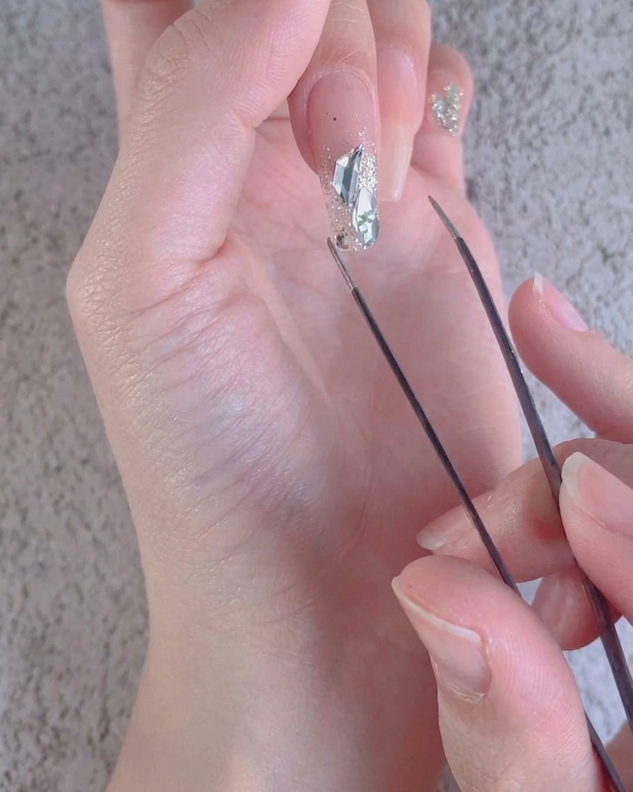 A close-up of tweezers adjusting small crushed diamond accents around larger stones on a nail.
