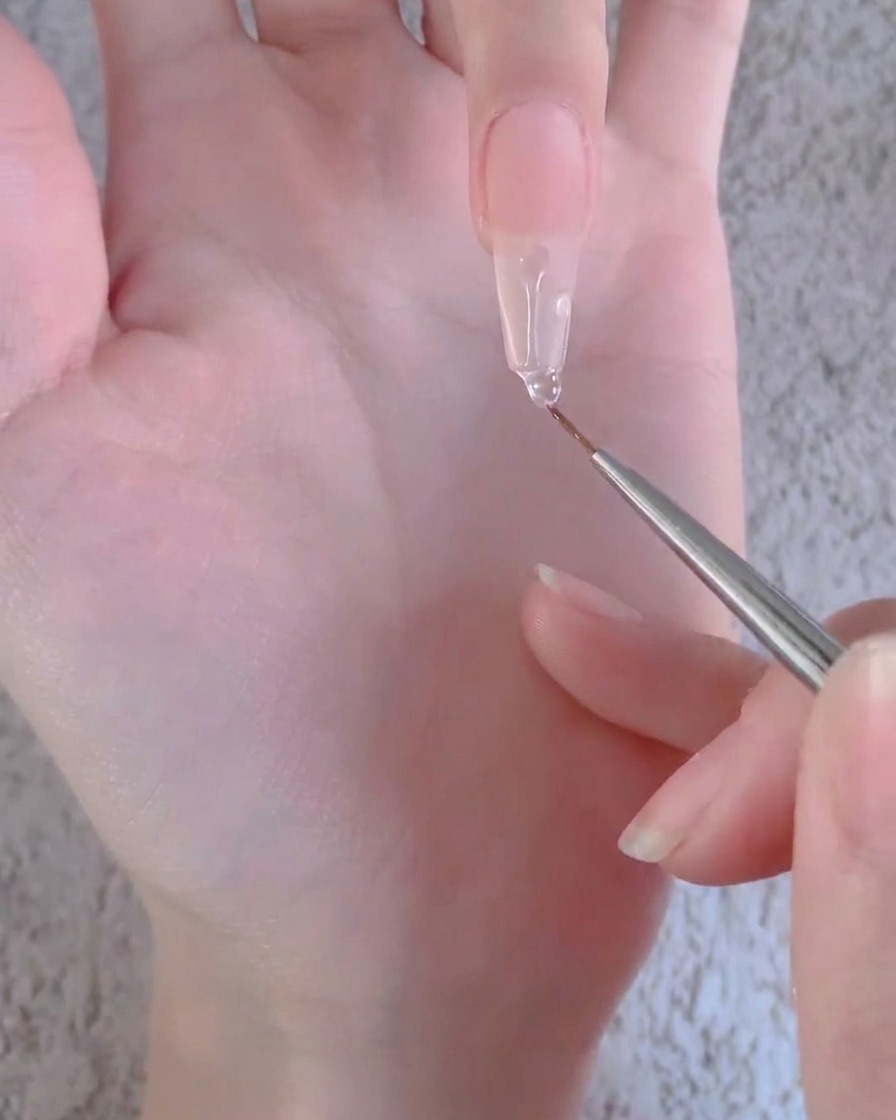 A liner brush pulling a clear gel bead downward along the length of a long artificial nail extension.