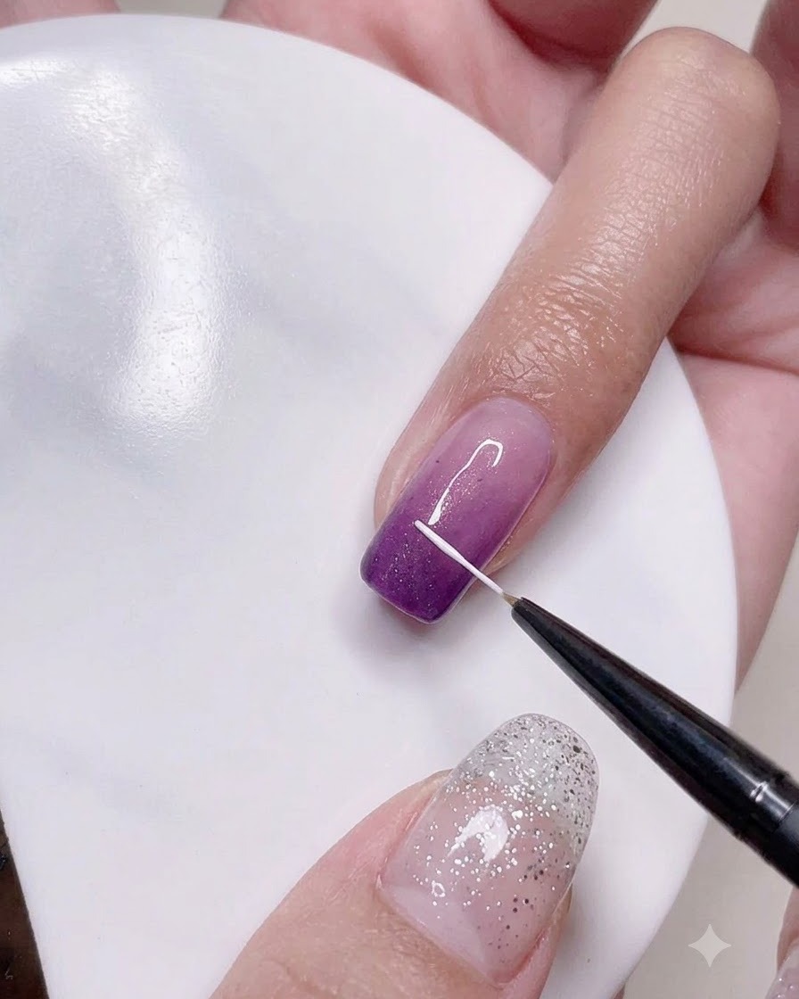 A fine liner brush drawing precise vertical white lines onto the purple ombre nail surface.