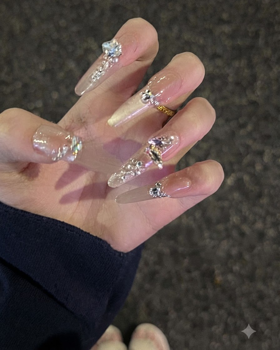 A close-up view of a hand with long nude rhinestone-studded nails curved slightly to show the profile.