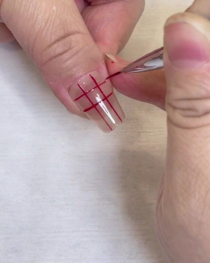 A close-up of a completed red grid pattern on a clear fingernail, drawn with a fine liner brush.