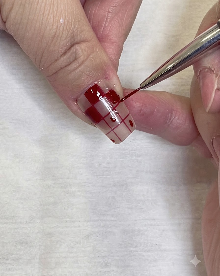 A detailed close-up of a fine nail art brush precisely applying red gel polish to the border of a checkered pattern on a nail.