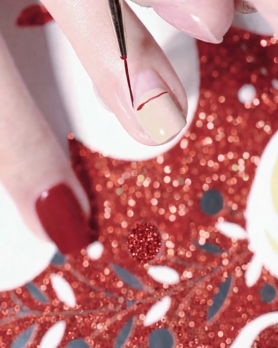 A close-up of a hand using a fine liner brush to draw a red curved line for a Santa hat on a nude-painted fingernail.