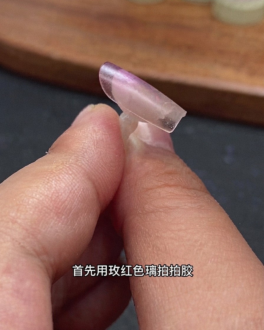 A close-up of a clear nail extension tip held by fingers with rose red glass patting gel applied at the back edge.