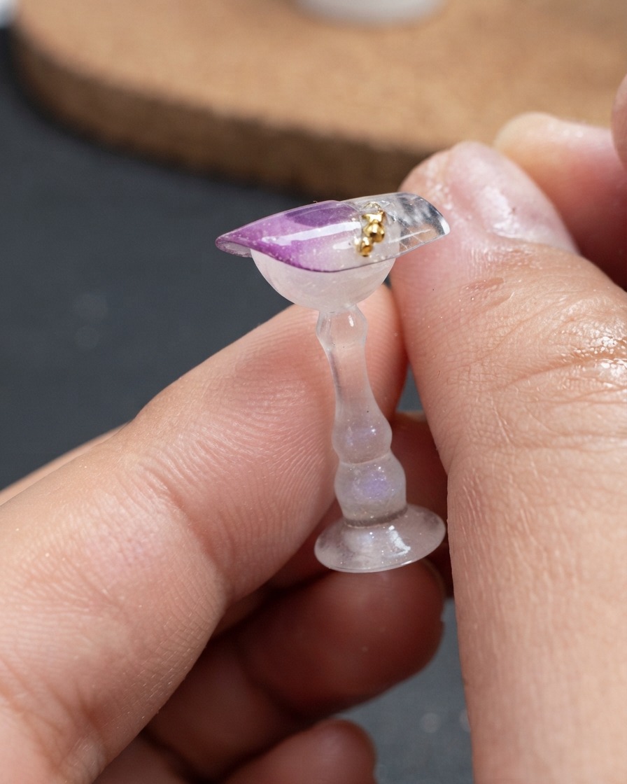 A close-up view of small gold beads being placed in a curved line on top of a decorated nail piece.