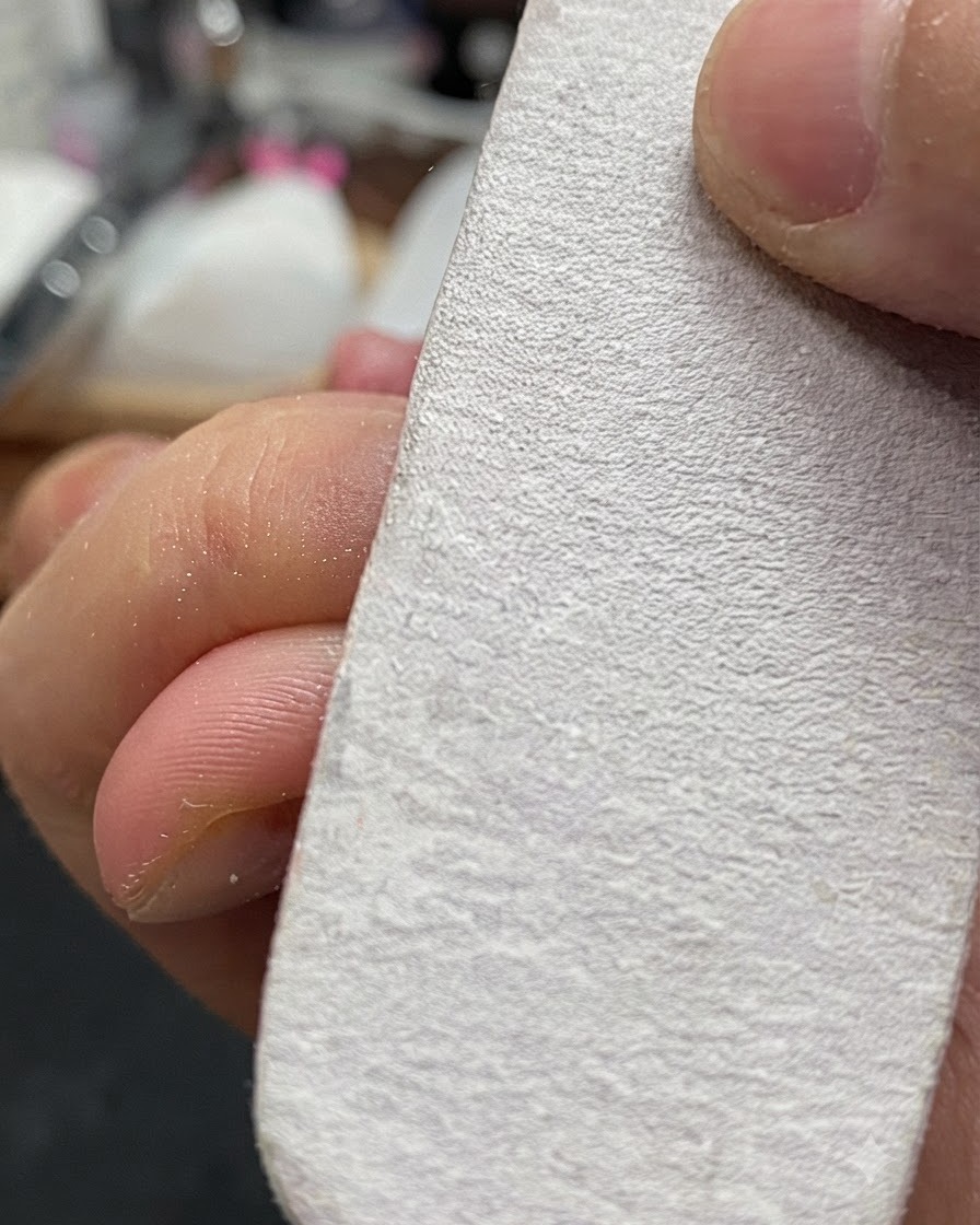 A hand holding a white nail buffer block lightly filing the surface of a natural fingernail.