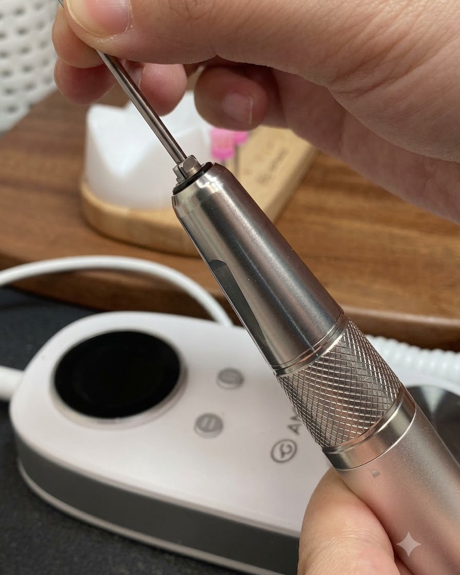 Hands inserting a long tapered metal drill bit into the handpiece of an electric manicure file.