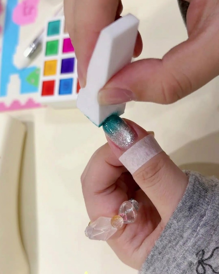 A white sponge block gently patting the tip of a manicured nail to build a dark teal gradient.