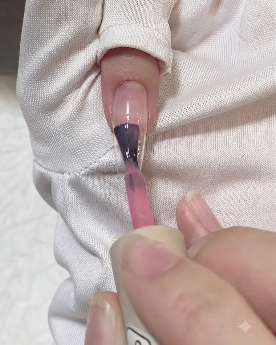 A brush applying a stroke of translucent pink gel down the center of a clear nail extension.