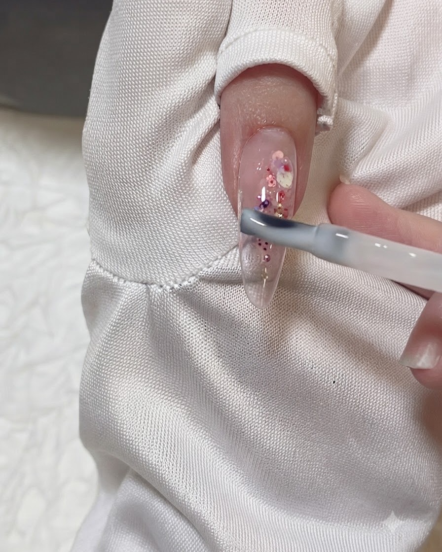 A gel brush applying a sheer milky gel layer over a clear almond nail decorated with pink and purple glitter flakes.