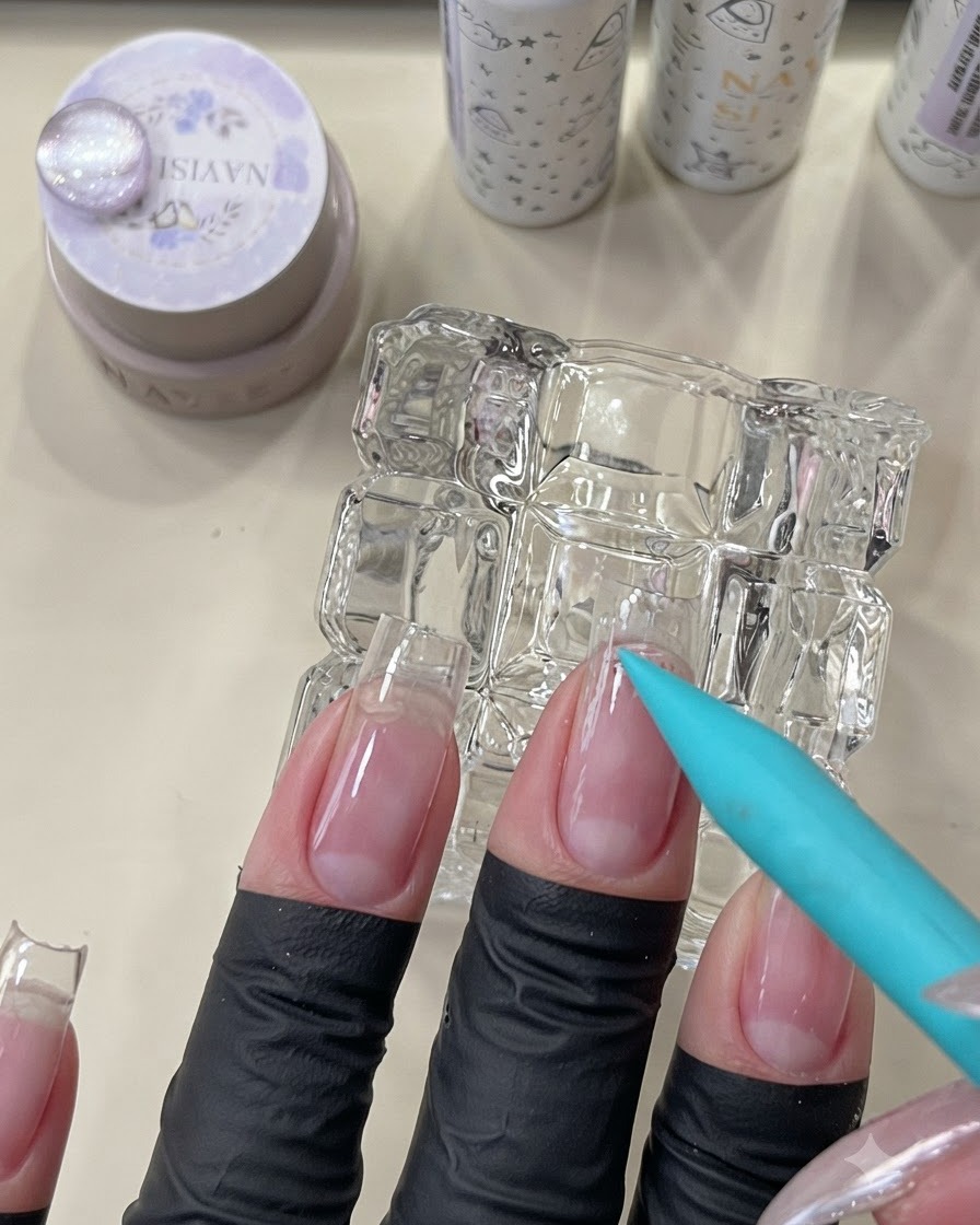 A blue brush applying a clear peel-off base coat to the cuticle area of a natural nail.