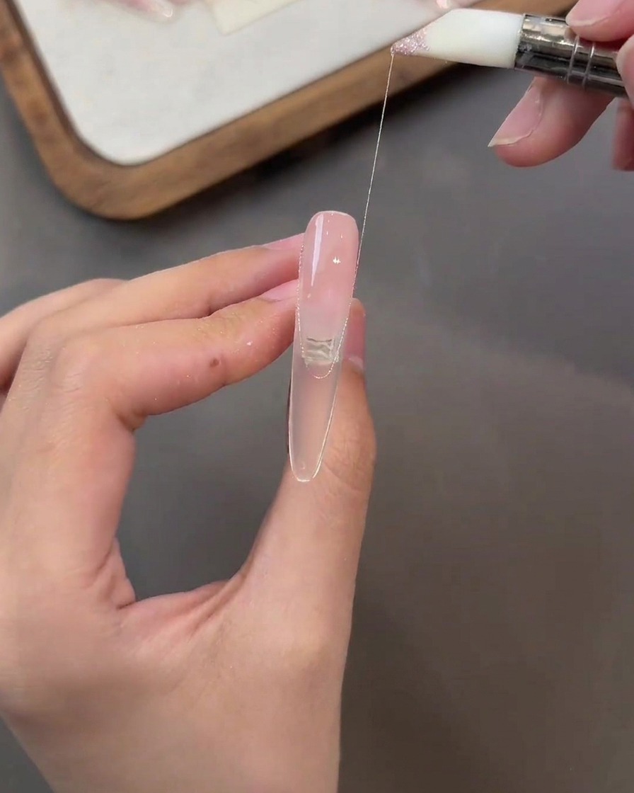 A brush pulling a thin strand of glitter pink gel across the surface of a nude-colored nail.