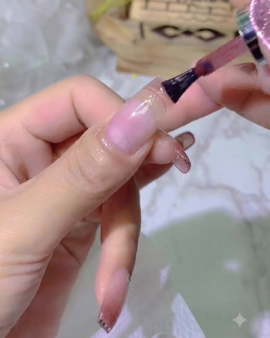 A close-up view of a brush applying jelly-toned color gel onto the nail plate, leaving a small gap near the cuticle.