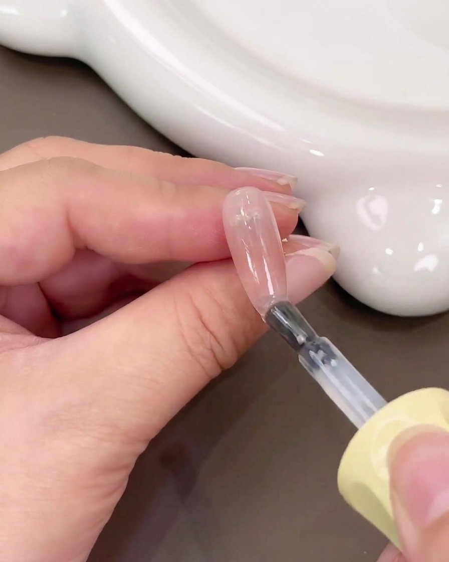 A brush applying clear base gel over a nail with a pink blush gradient.