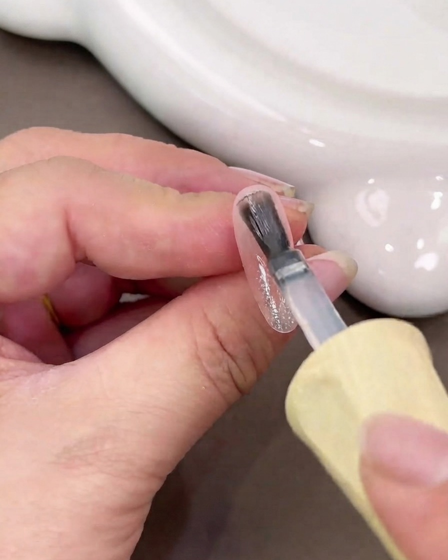 A nail brush applying a wet layer of clear base gel onto a glossy nail surface.