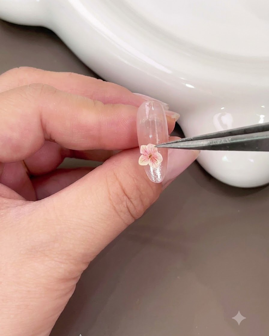 A pair of tweezers placing a small pink dried flower onto the lower portion of a glossy nail.