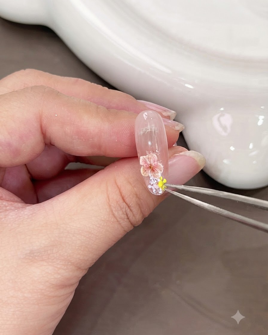 A pair of tweezers placing a small yellow dried flower next to the pink flower on the nail.