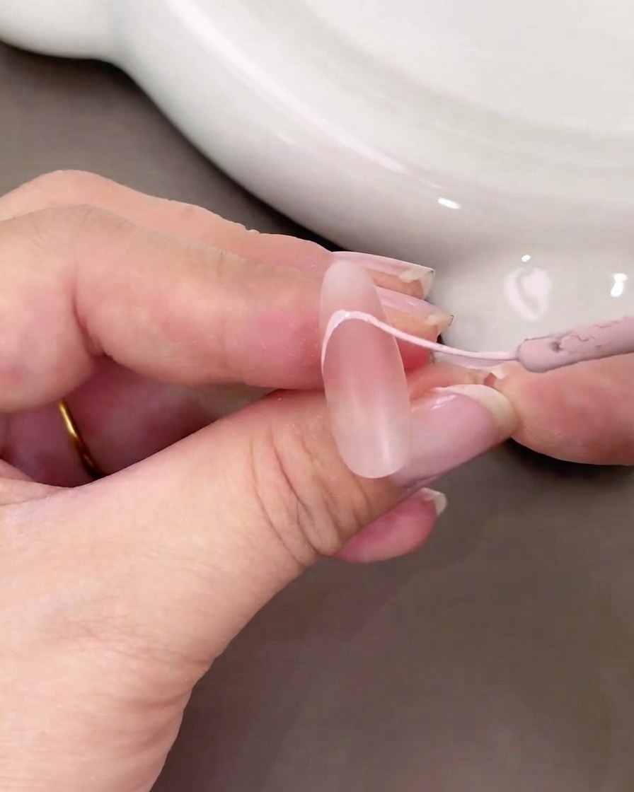 A close-up of a hand using a fine brush to paint a light pink French tip curve on a nude nail.