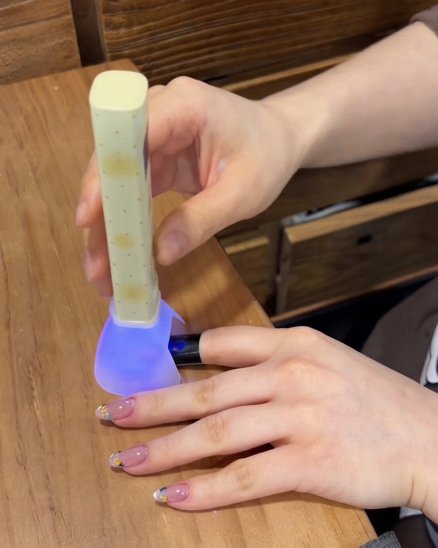 A portable purple UV/LED nail lamp curing the gel on a fingernail while other manicured fingers rest on the table.